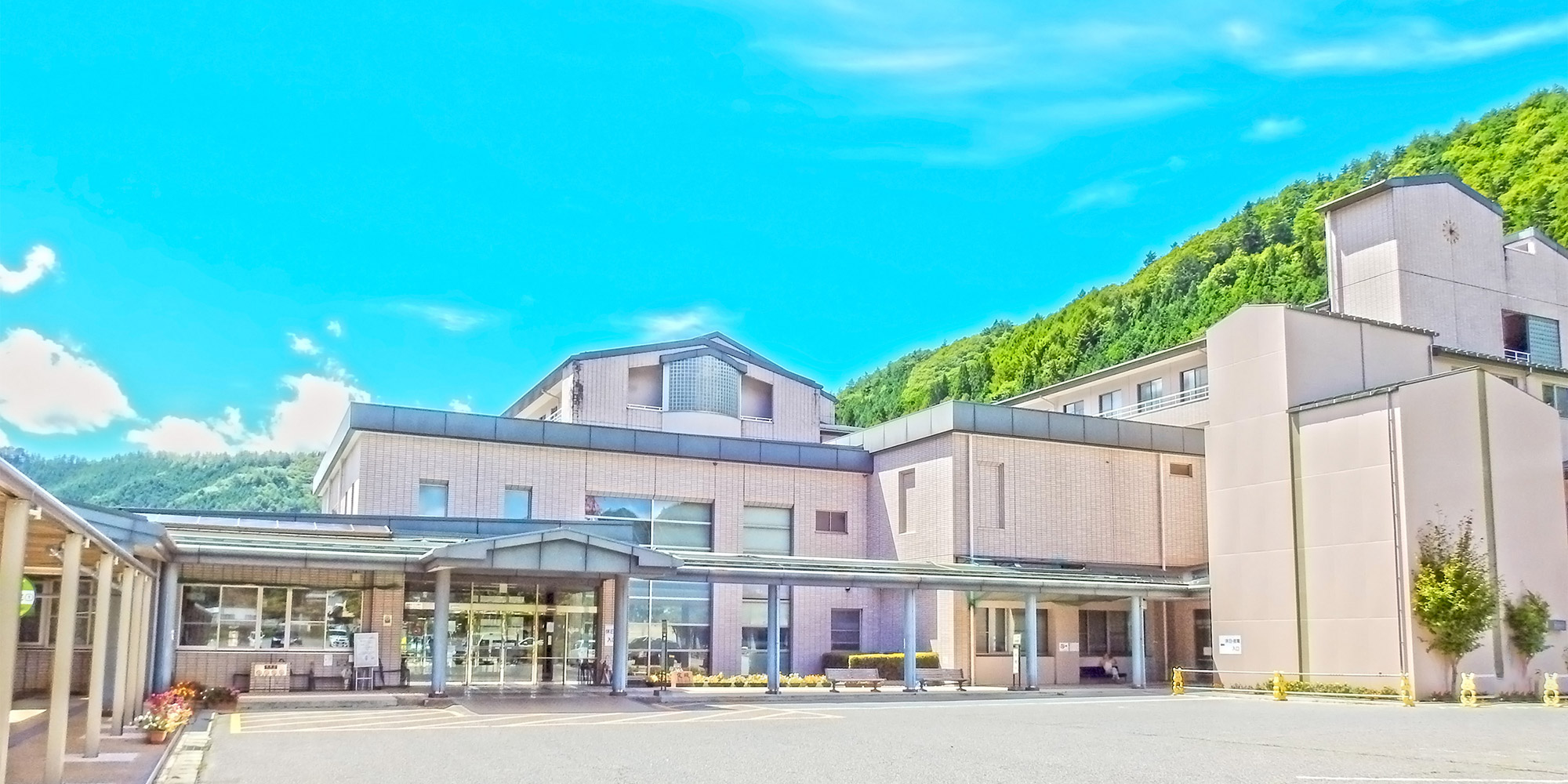 山々に囲まれた介護施設の建物外観。モダンな平屋建て構造で、広い駐車場が備わっている。